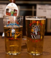 Two pints of beautiful looking beer, one partially drunk, sit atop the beer festival bar, with a slightly out of focus hand pump behind them.